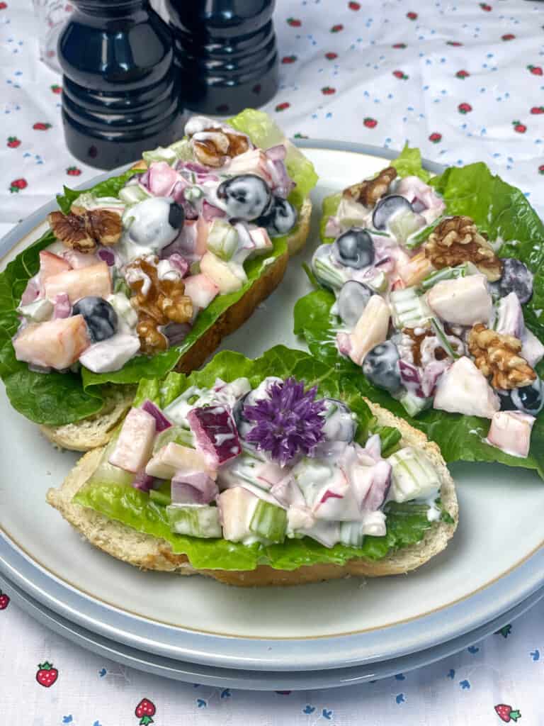 A light blue plate with two slices of crusty bread with lettuce and vegan Waldorf salad filling, salt and pepper shakers to side, strawberry images on a white tablecloth background.