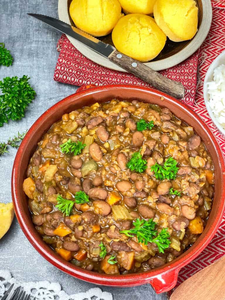 red beans stew served in red serving platter, with plate of cornbread muffins in background, bowl or rice to side, parsley on grey work surface, and red tea towels.