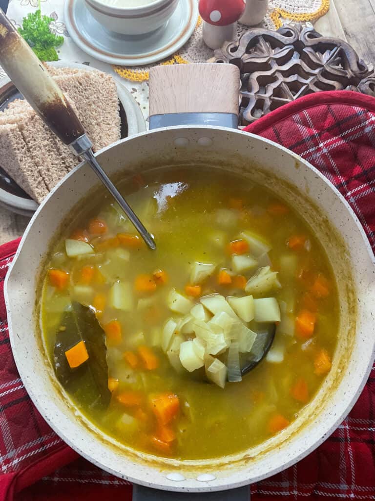 Scottish potato soup cooked and ready to serve with wooden handled ladle, bread slices on plate to side, soup bowls to side and red tartan towel to side.