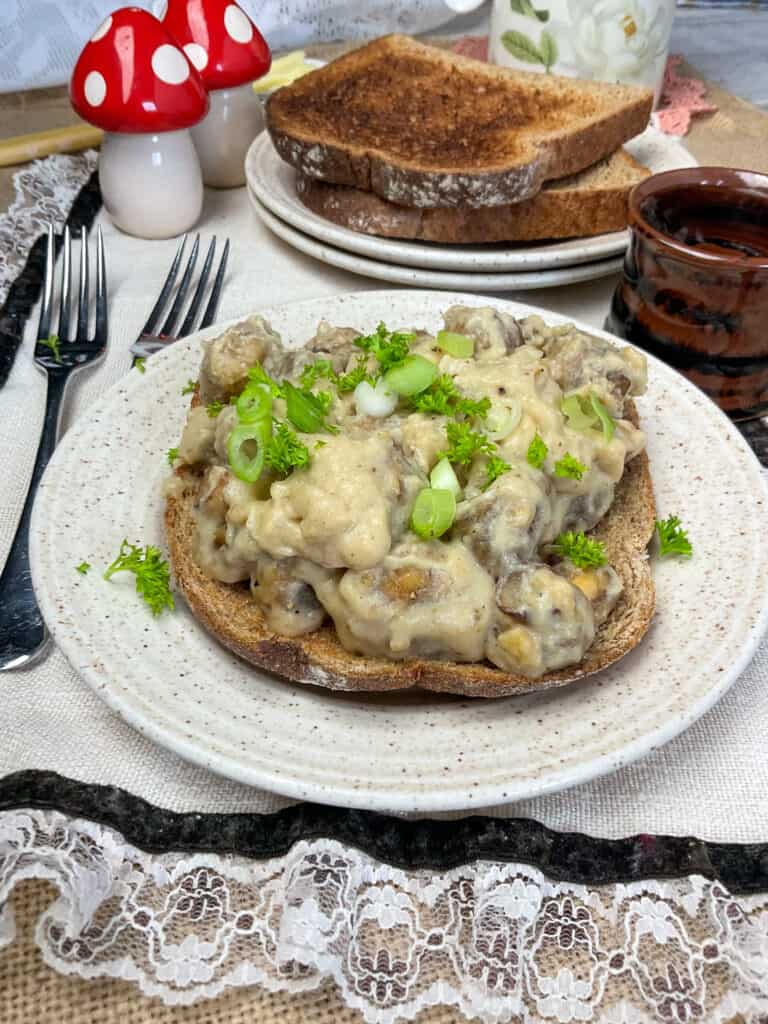 creamed mushrooms on toast served on white speckled plate with cutlery to side, toasted bread slices in background, small brown cup to side and mushroom shaped salt and pepper shakers, frilly cream place mat underneath.