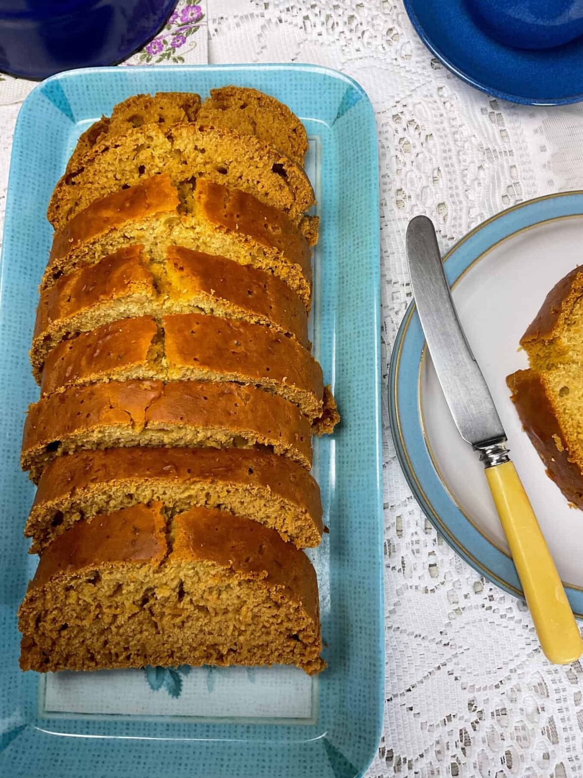Golden syrup loaf baked and sliced, on a blue serving tray, blue rimmed plate to side with yellow handled butter knife, blue cups to side and a white net table cloth underneath.