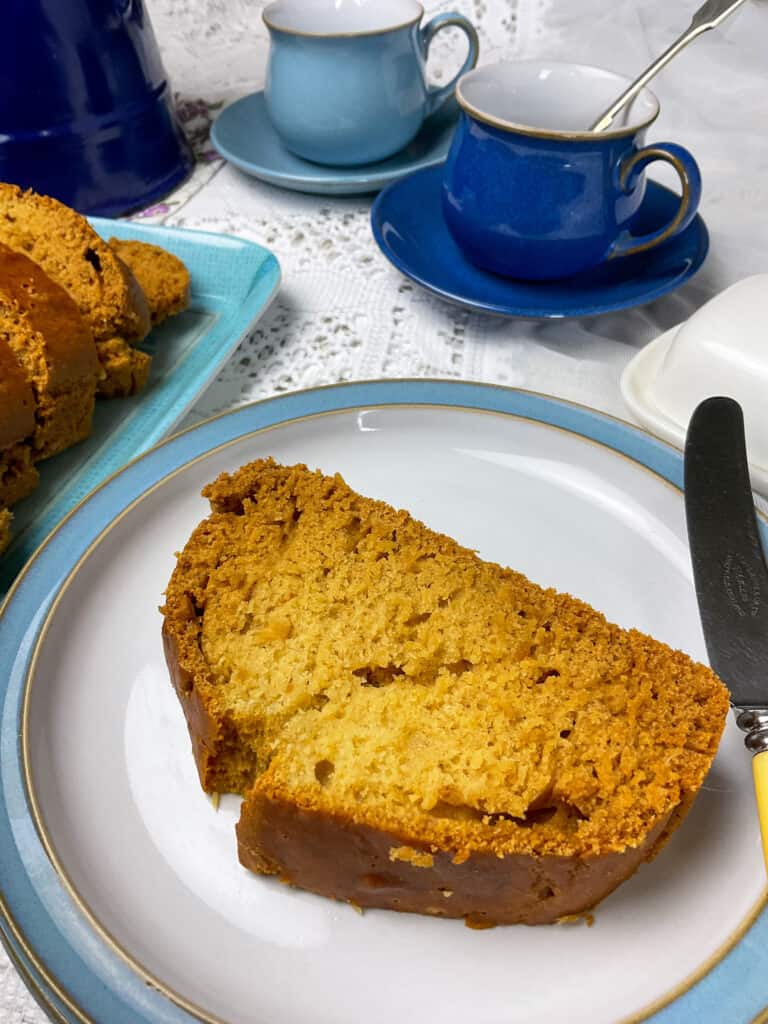 slice of syrup loaf on blue rimmed plate with two blue toned cups and saucers clearly in background, blue coffee pot to side and blue tray with rest of syrup loaf slices, white net table cloth. 