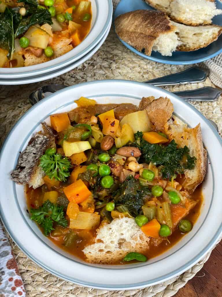 a bowl of zuppa di cavolo served with blue side plate of ripped up crusty bread and second bowl of soup to side, raffia placemat background.