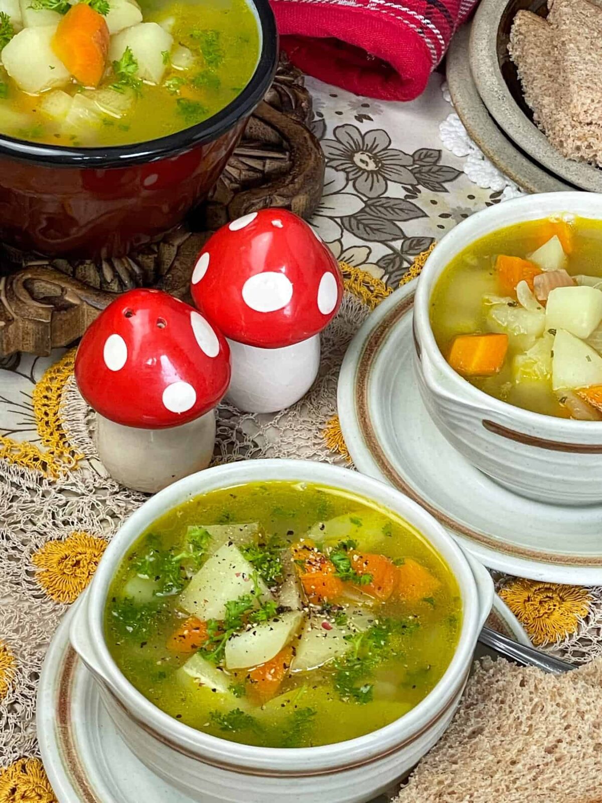 Table setting with two small bowls of Scottish potato soup with brown saucepan filled with soup in background, red and white mushroom shaped salt and pepper shakers with flower tea towel background.