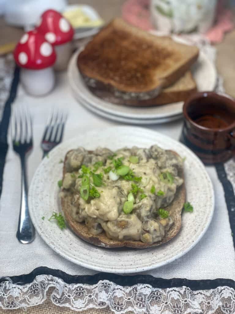 table setting with plate of creamed mushrooms on toast with second plate of toasted bread in background with small brown  cup, forks to side and ruffled vintage table mat.
