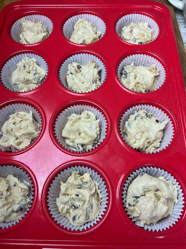 Granny cake batter divided between a red 12 cup muffin tray lined with paper cases.