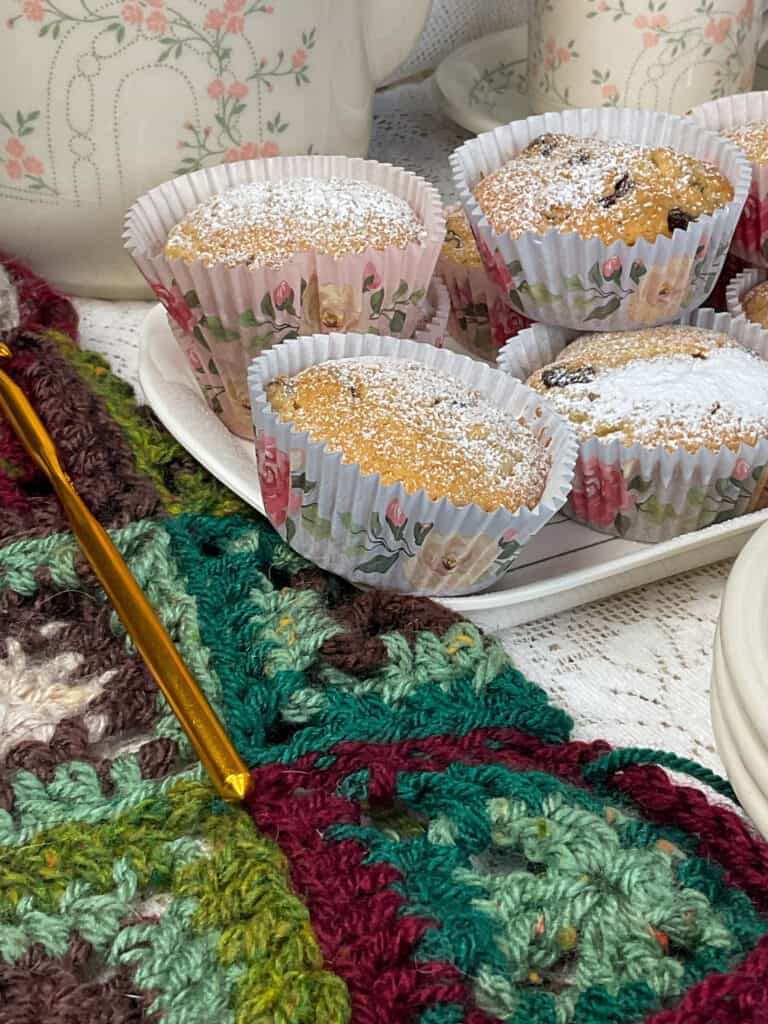 A side view of a tray of Granny cakes, with green and brown coloured shades of wool crochet to side and a gold crochet hook. Teapot and cup and saucer in background, with white doily table cloth.
