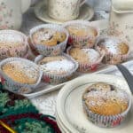 A small plate with a Granny cake and small cake knife to side, tray of more Granny cakes in background along with matching flower patterned teapot, cup and saucer and milk jug, green and brown crochet squares with gold crochet hook to side, white doily tablecloth.