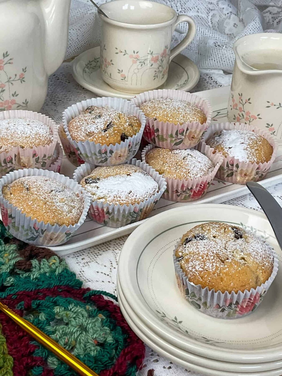 A small plate with a Granny cake and small cake knife to side, tray of more Granny cakes in background along with matching flower patterned teapot, cup and saucer and milk jug, green and brown crochet squares with gold crochet hook to side, white doily tablecloth.