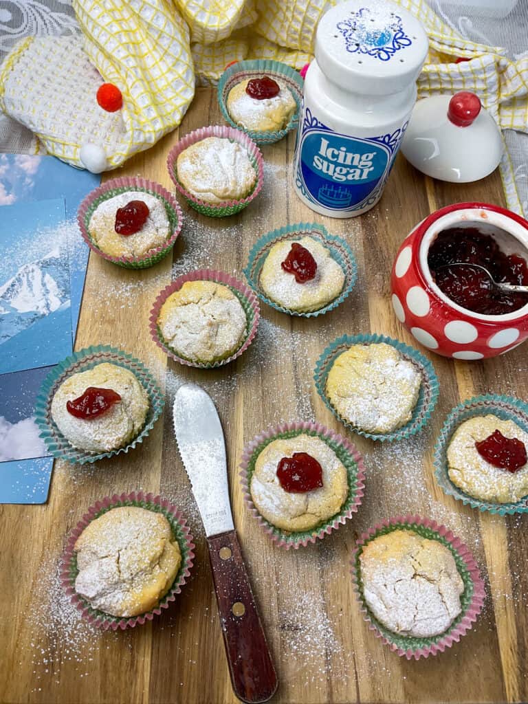 Swiss cakes decorated, with red and white spotty jam pot to side, blue and white icing sugar decanter to side, wooden handled spatula on wooden board, yellow tea towel background, photos of mountains and blue sky to side.