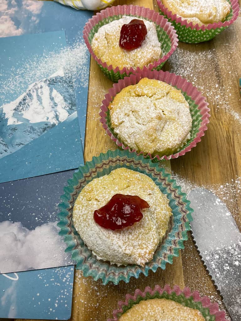 a row of Swiss cakes decorated with fruit jam and dusted with icing sugar, photos of snowy mountains and blue skies to side, on wooden chopping board.