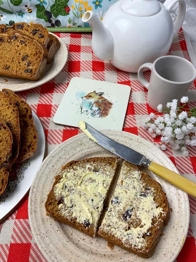 table set for malt loaf and tea pot and cup, tea mat with image of fox, and small jug of white flowers to side, red and white check tablecloth.