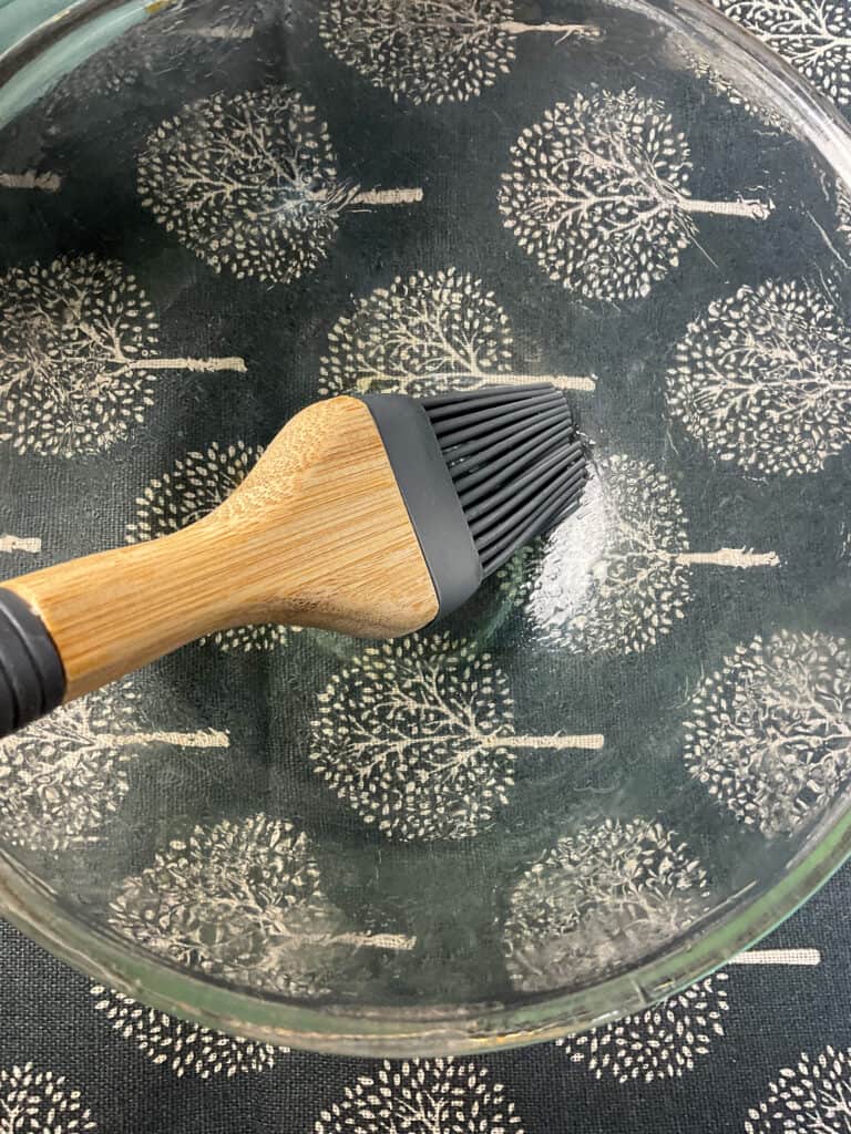 Glass pudding bowl being greased with oil with a pastry brush, grey tree lined table mat underneath.