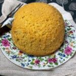 Close up of steamed cornbread on flower patterned plate with cutting knife and fork to side, and rustic cream coloured table mat.