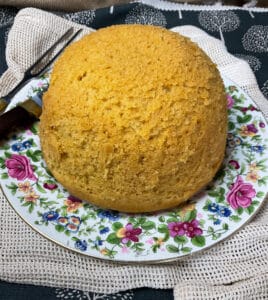 Close up of steamed cornbread on flower patterned plate with cutting knife and fork to side, and rustic cream coloured table mat.