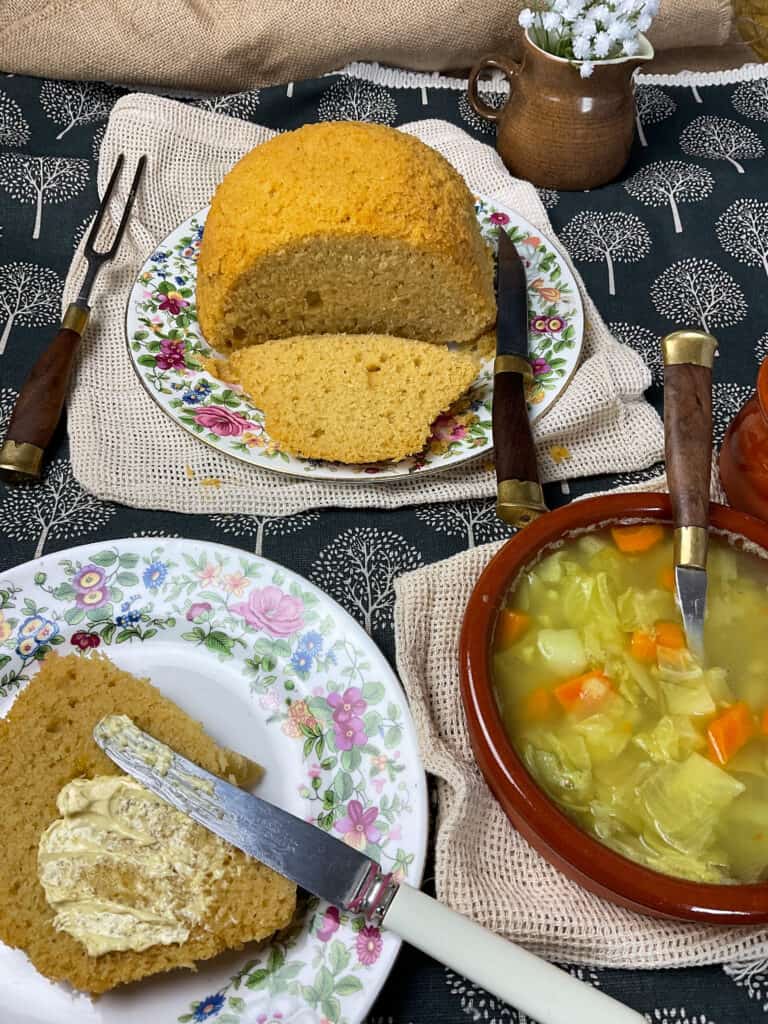 cornbread served on flower patterned plate with a slice cut and a second plate with a slice of cornbread buttered with knife, brown bowl of soup to side and small brown jug in background with white flours, blue tablecloth with white trees background.