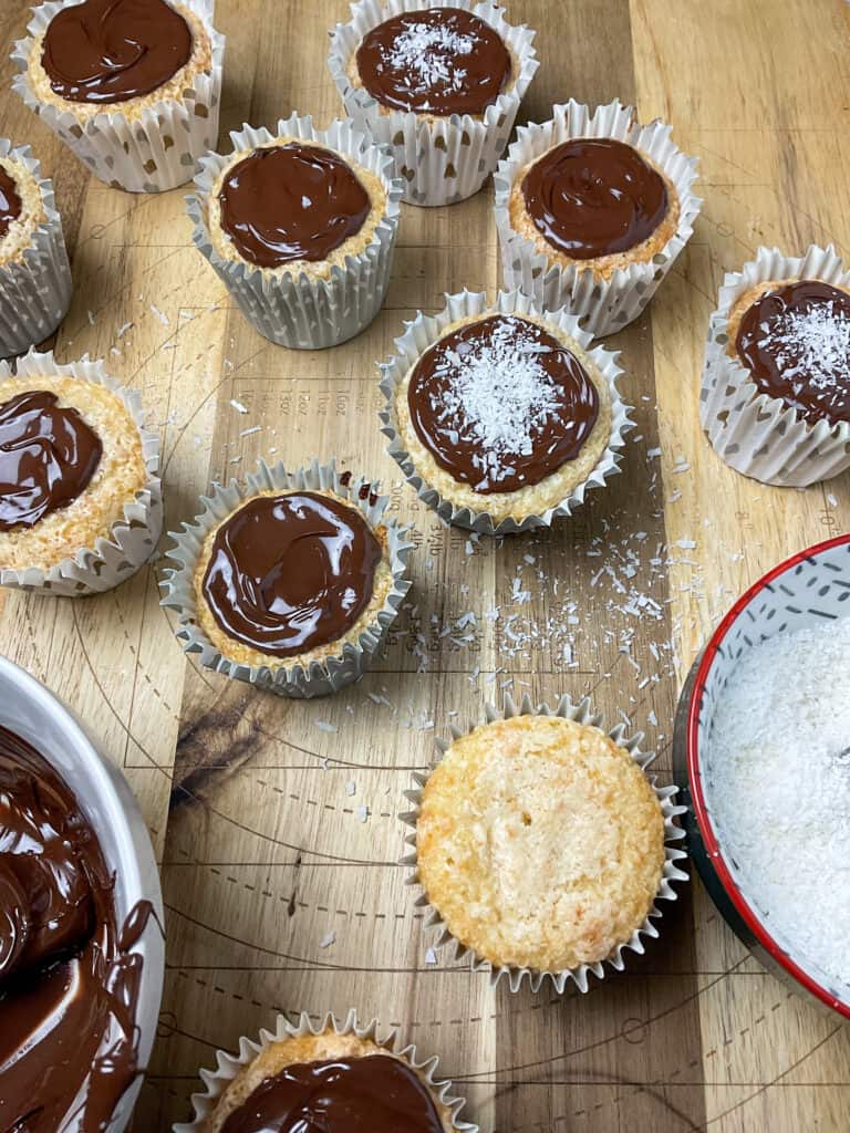 coconut buns decorated with melted chocolate and desiccated coconut, bowl of melted chocolate to side, and bowl of coconut to side, wooden pastry board background.