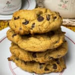 a pile of 4 chocolate chip cookies on a small plate with pink flower design, teapot and cup and saucer to background with yellow patterned table cloth.