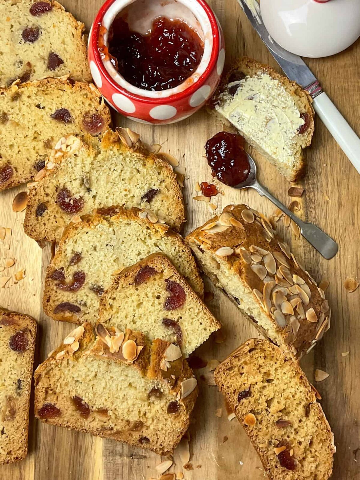 Cherry loaf on wooden board, cut into slices with jam pot to background, jam spoon and butter knife to side.