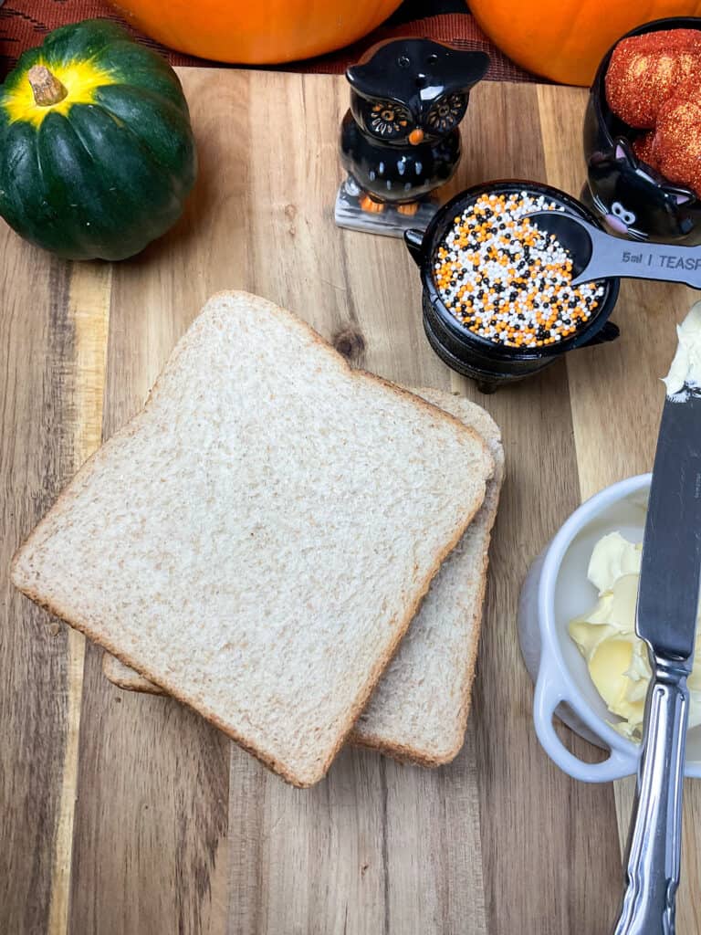 slices of bread, dish of cake sprinkles, small green pumpkin and small black and orange owl ornament, with butter dish and knife to side, on wooden chopping board.