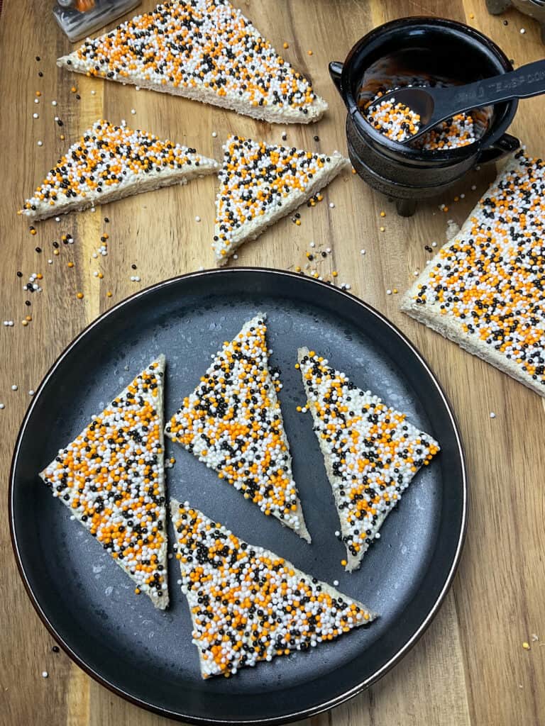 black plate with triangles of fairy bread and more sliced fairy bread to side with small cauldron holding cake sprinkles, on chopping board.