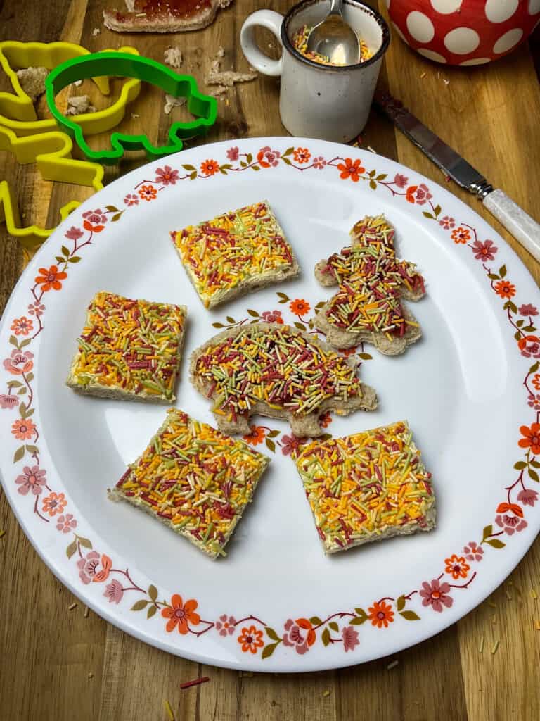 a flower patterned rimmed plate with cookie cutter bread shapes covered with cake sprinkles, cookie cutters to side, small coffee cup filled with sprinkles, wooden board background.