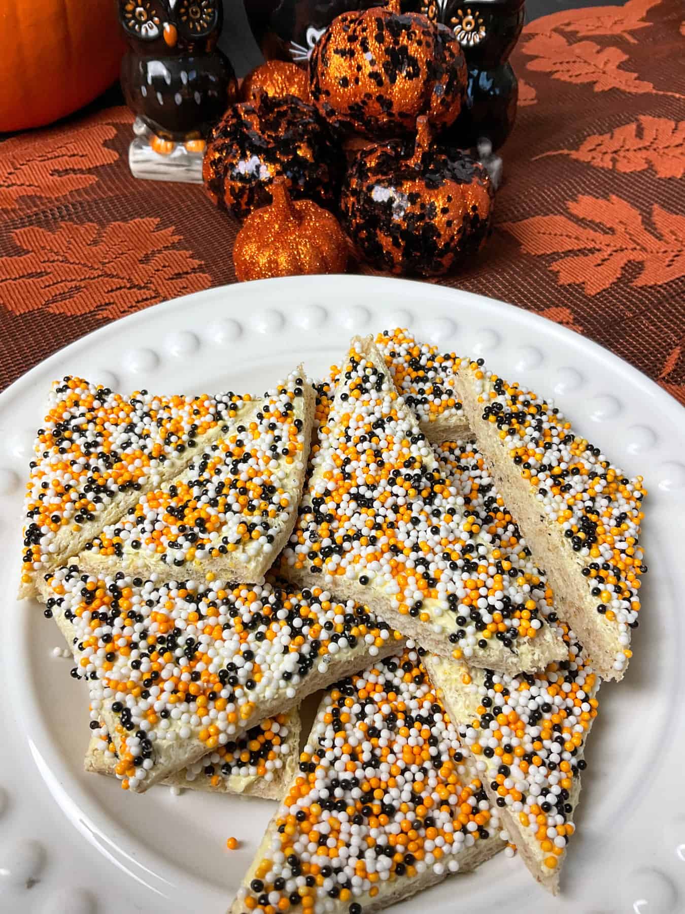 A white plate of Fairy bread with white, black and orange sprinkles, pumpkin ornaments in background