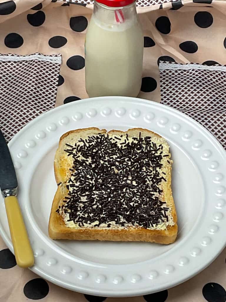 a slice of Hagelslag on a white plate with butter knife to side, bottle of milk with red lid and straw in background, brown spotty tablecloth background.