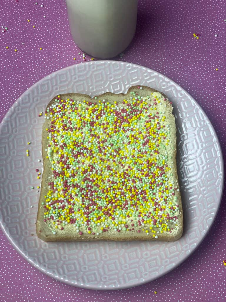 a pink plate with slice of Fairy bread, milk bottle in background and pink sparkly background mat.