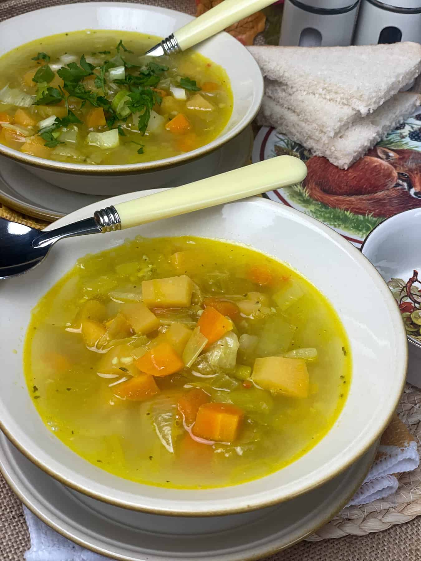 Close up bowl of vegetable soup with yellow handled spoon and second bowl in background, slices of bread to side on a mat with fox image.