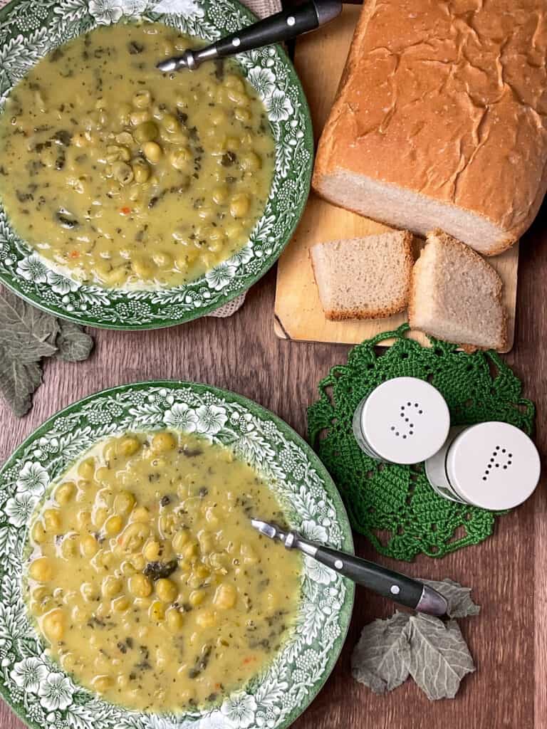 Ariel photo of two bowls of pea soup with green handled spoons and loaf of bread to side on small chopping board, salt and pepper shakers, dried mint leafs decor and dark wooden background.