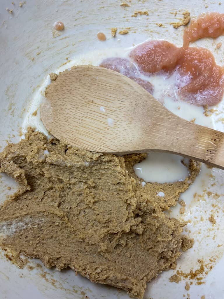 Ingredients creamed together and apple sauce and milk added to mixing bowl with wooden spoon.