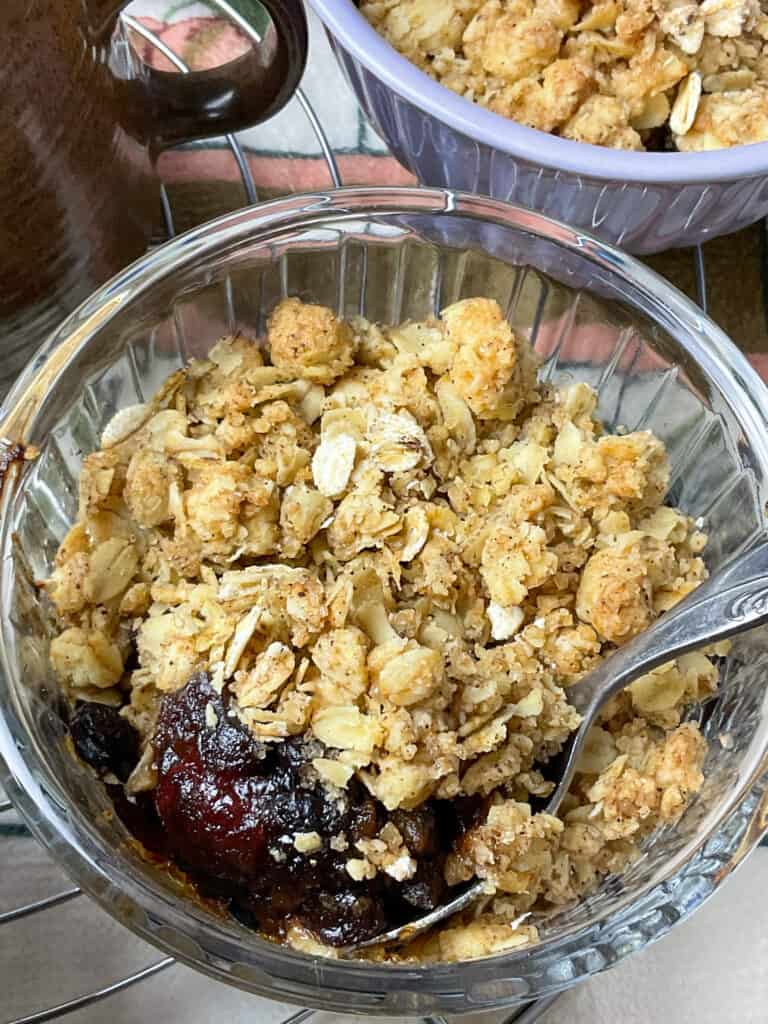 close up of dried fruit crumble with a spoon scooping up the stewed fruits, brown jug handle to side and second pudding to side.