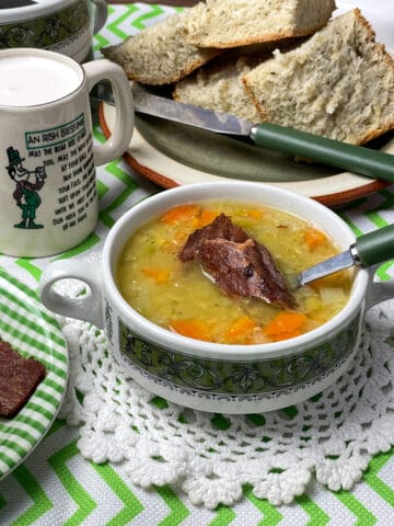A bowl of vegan pea and 'ham' soup in green patterned bowl, with green handled spoons, white doily underneath, bread slices on green plate in background, small jug with cream and green and white striped table cloth.