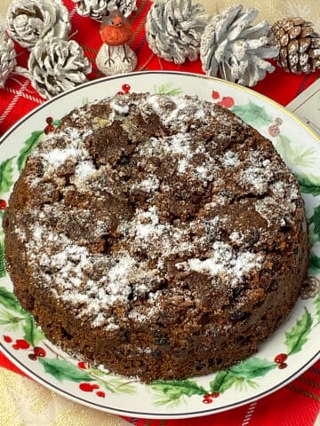 close up of vegan microwave clootie dumpling on festive leaves decorated plate, white frosted pinecones to background, small robin cake ornament to side.