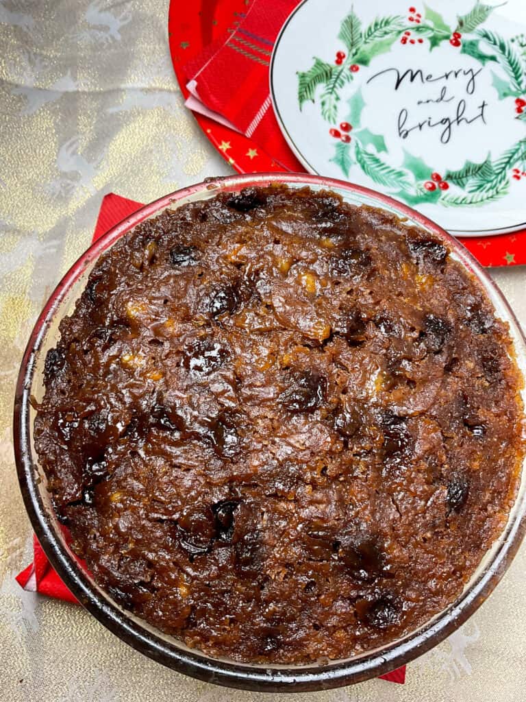 cooked Christmas pudding still in pudding basin on dinner table with serving festive plate to side and red circular tray.