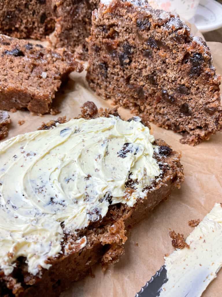 A close up of a slice of clootie spread with vegan butter, slices of clootie in background, wooden chopping board background.