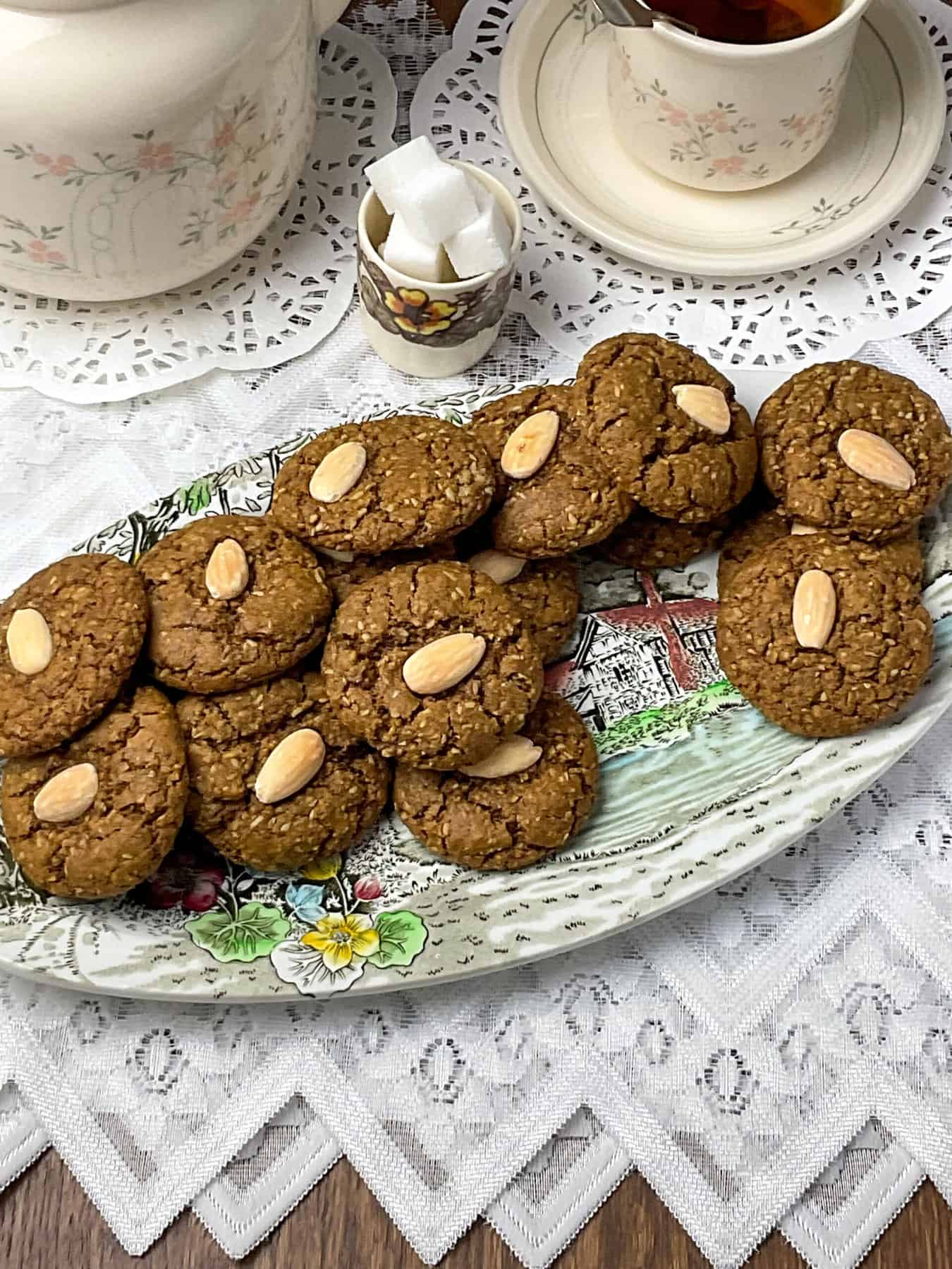 An oval serving plate filled with parkin biscuits, cream coloured flower patterned tea pot in background with matching cup and saucer, small sugar cube dish, and white doily tablecloth with brown wood background.