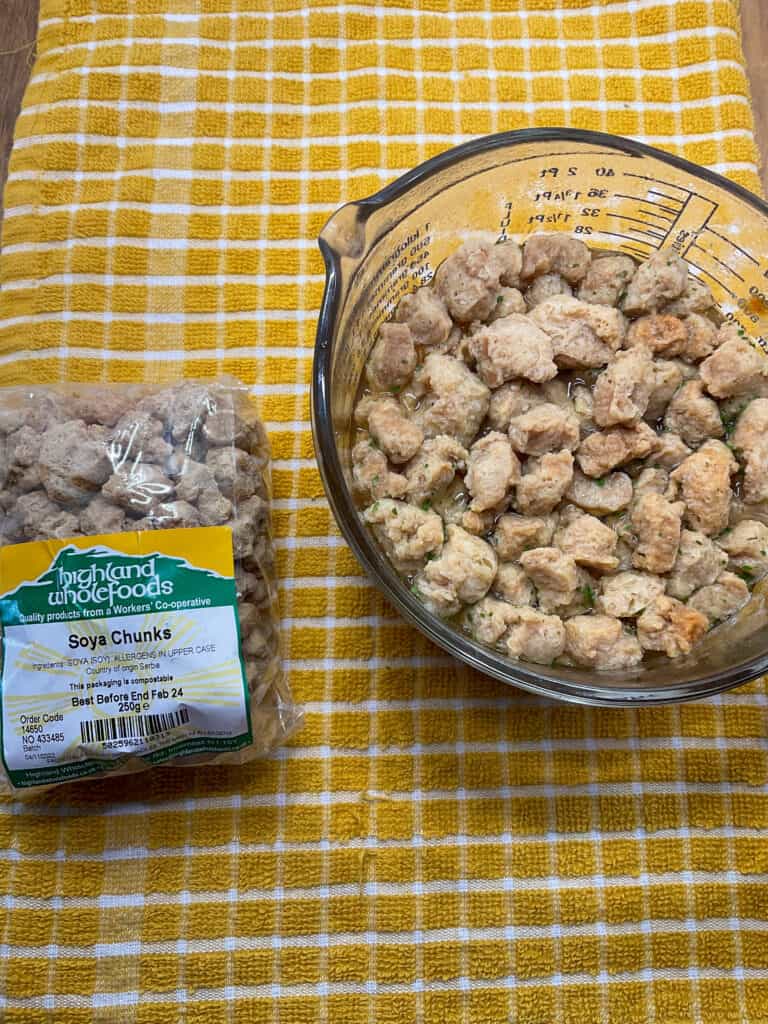 soya chunks in glass bowl soaking in veggie stock, packet of soya chunks to side, yellow tea towel background.