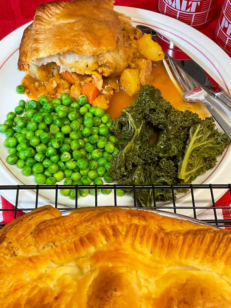 slice of vegan meat and potato pie served on white plate with red rim, and sides of peas and kale with gravy, whole pie to front sitting on wire rack, red table mat background.
