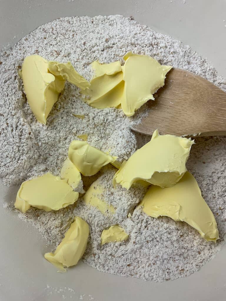 vegan margarine added to flour mixture into bowl with wooden spoon to side.