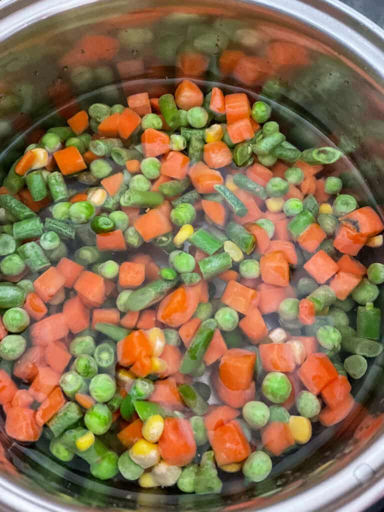 Frozen mixed vegetables in a sliver saucepan with water covering ready to cook.