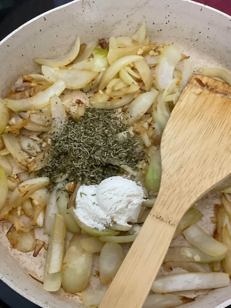 flour and herbs added to cooked onions and garlic in fry pan with wooden spatula.