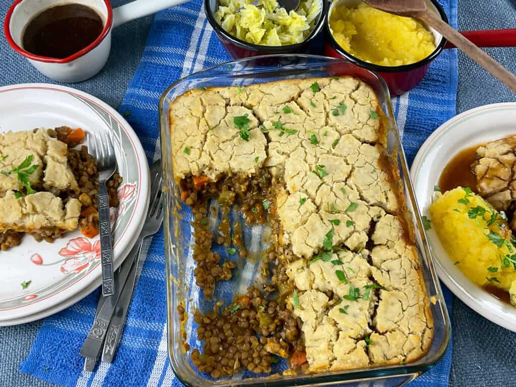 Teviotdale pie in casserole dish with two portions removed, two dinner plates with pie to side, and pots of veggie sides and gravy to background.