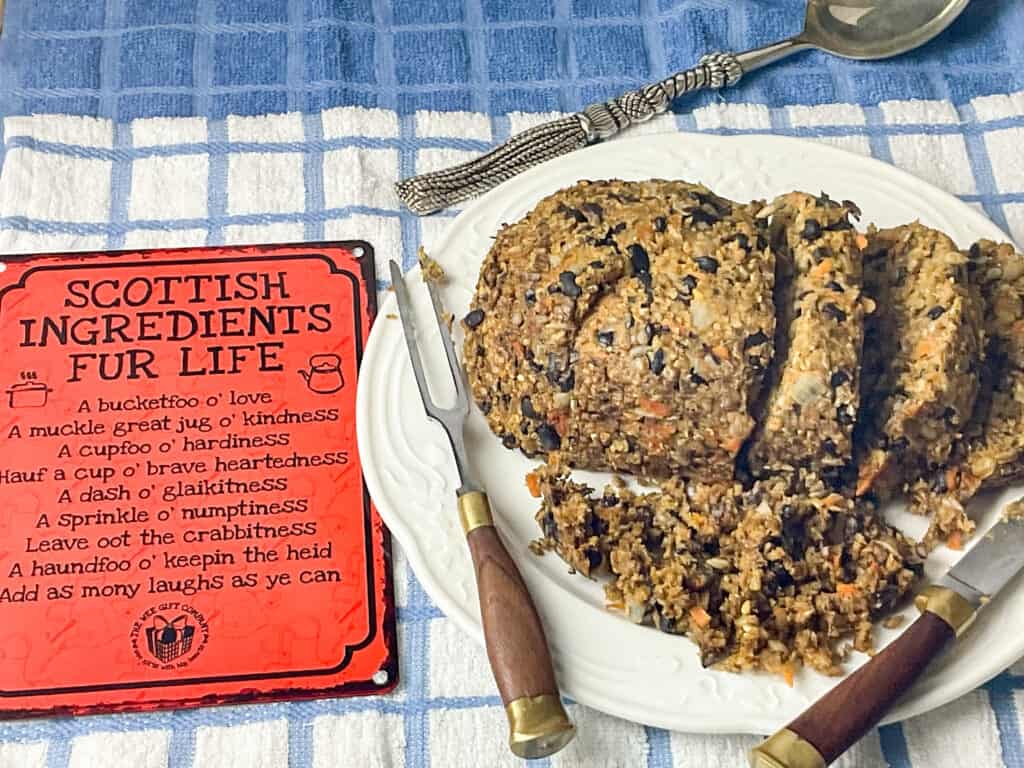 vegan haggis served on white serving plate with some of the haggis crumbled up, brown handled carving knife and fork on plate, red Scottish sign to side, silver serving spoon to background, and blue and white check table cloth.