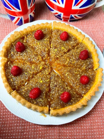 A white serving plate with a whole treacle tart sliced into 8 pieces, each slice with a raspberry garnish, British flag teapot with matching cup in background, red tea cloth background.