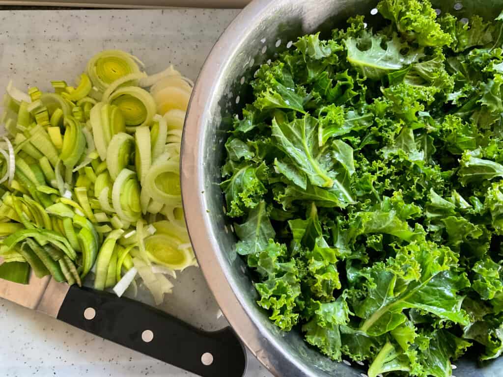 A white chopping board with black handled knife with bowl of prepared kale and sliced leeks.