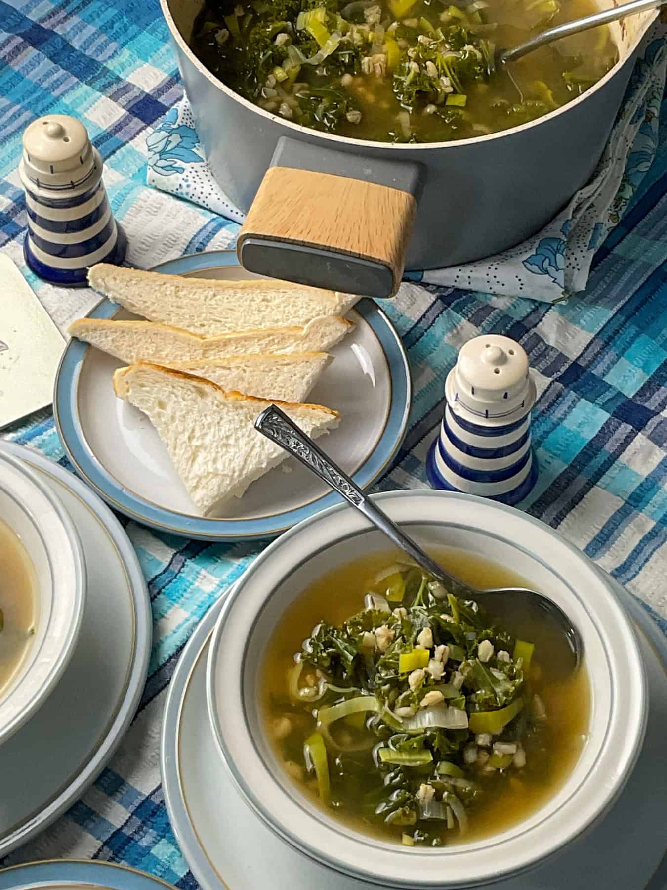 A table setting with a bowl of barley kale broth, small plate with bread slices, blue and white salt and pepper shakers, soup pot in background and a blue stripe table cloth.