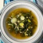 A close up bowl of barley kale broth served in a blue rimmed bowl, and a blue and white checked tablecloth background.