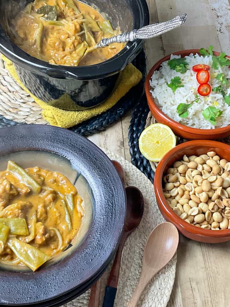 A dark brown bowl with a serving of Thai red curry, two smaller light brown bowls with rice and one with peanuts to side, three wooden spoons to side and the slow cooker pot in background with silver ladle.